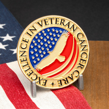 Gold challenge coin with the text Excellence in Veteran Cancer Care featuring an eagle silhouette over an American flag design.
