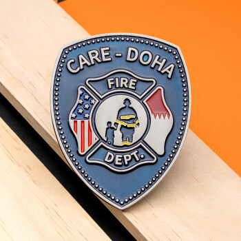 Shield-shaped lapel pin for Care-Doha Fire Dept featuring a maltese cross, American and Qatari flags, and a firefighter silhouette.