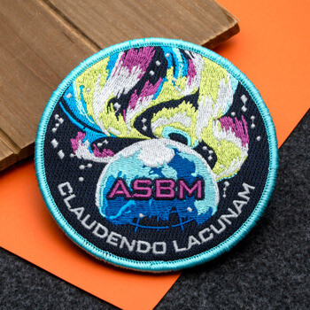 Circular embroidered patch with ASBM text over a globe and colorful aurora design, bordered by the words Claudendo Lacunam.