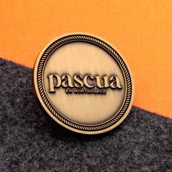 Circular antique gold challenge coin featuring the Pascua brand name with a rope border design, resting on a grey and orange background.