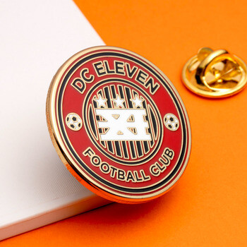 Round red and gold DC Eleven Football Club enamel lapel pin shown with a butterfly clutch back against an orange background.